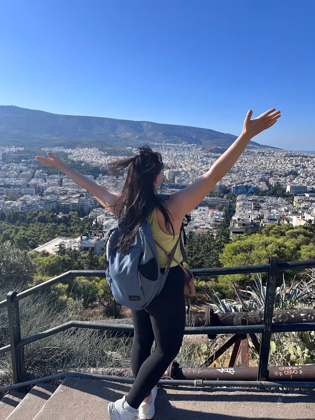 Walker with arms raised at an Athens hills viewpoint, Mount Hymettus rising across the city behind her