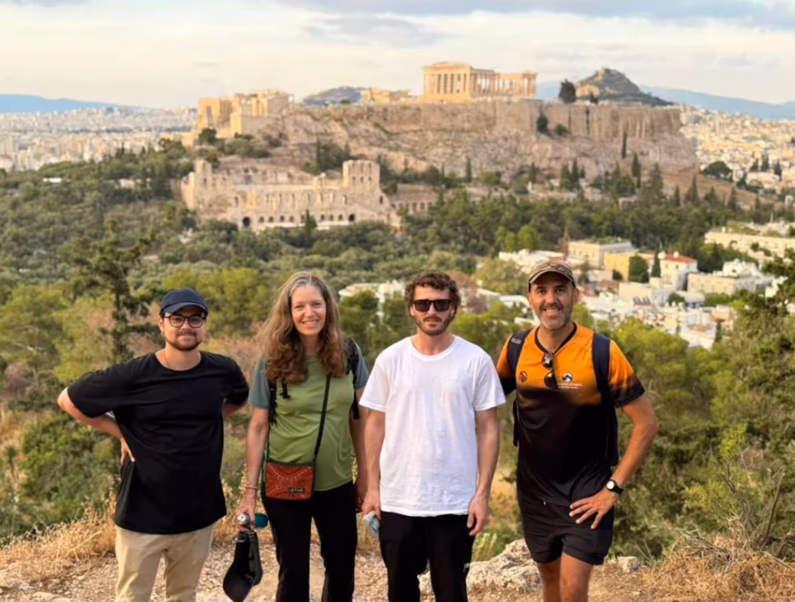 Tour group with George in the orange Ultimate Athens shirt at an Athens hilltop viewpoint, the Acropolis behind them