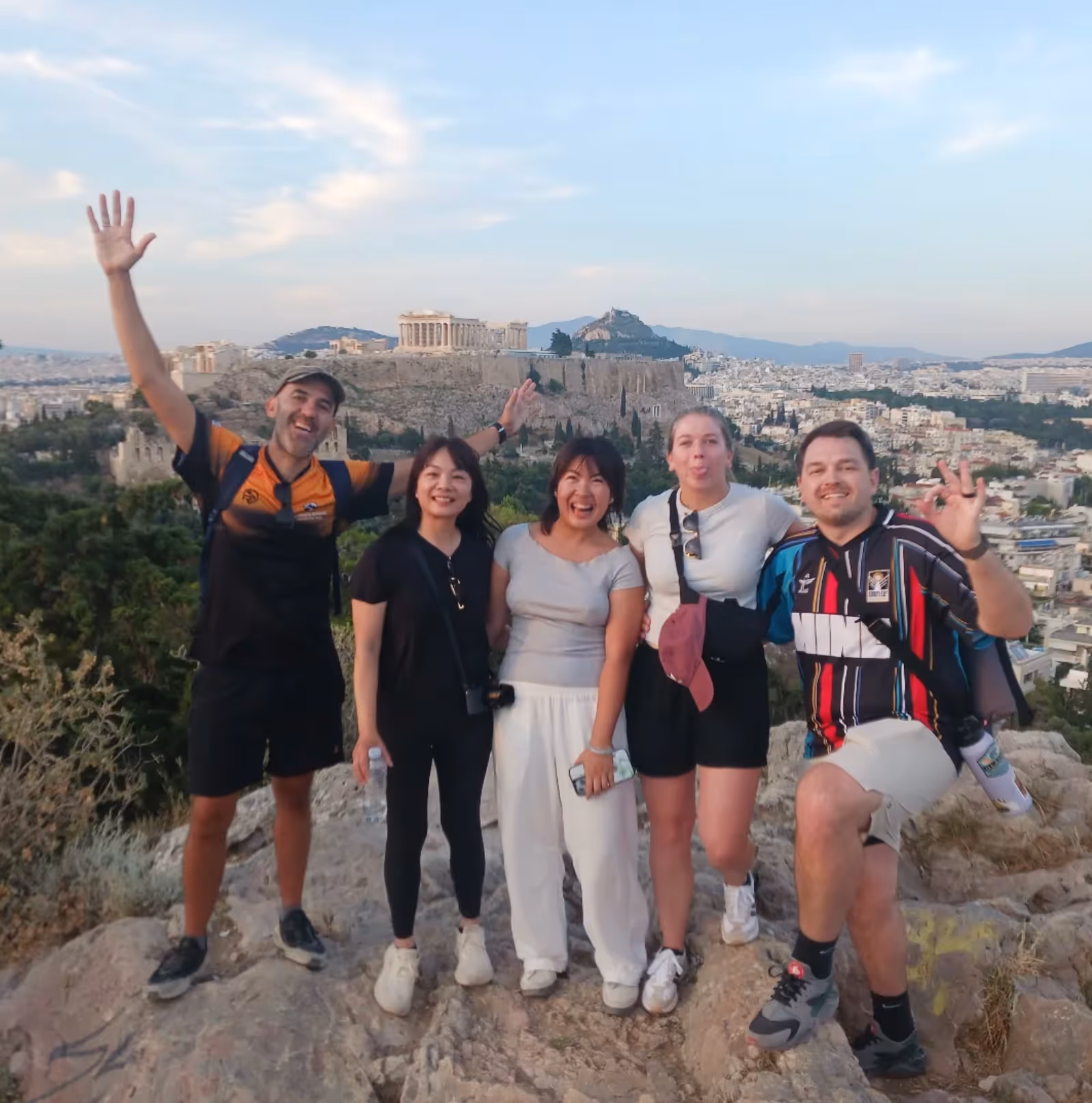 Group of guests celebrating with arms raised above Athens, the Parthenon and Lycabettus Hill on the skyline behind them